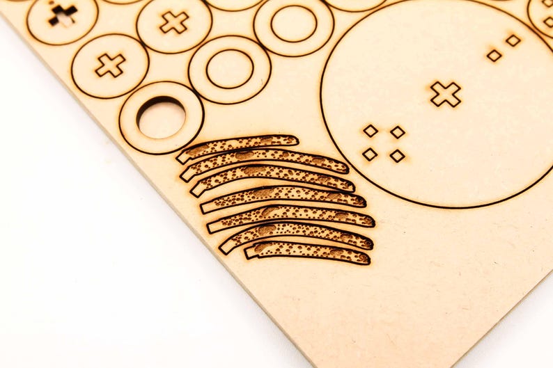 May include: A close-up of a wooden sheet with laser-cut shapes. The shapes include circles, plus signs, and curved lines with dotted patterns. The wood has a light brown color and a smooth texture. The design suggests a craft or DIY project.