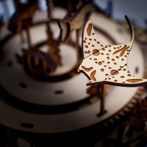 May include: A wooden laser-cut model of a manta ray with a spotted pattern, set against a blurred background of a clock mechanism. The ray is light-colored with brown details, showcasing the intricate design.