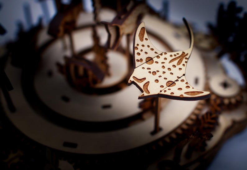 May include: A wooden laser-cut model of a manta ray with a spotted pattern, set against a blurred background of a clock mechanism. The ray is light-colored with brown details, showcasing the intricate design.