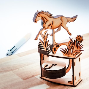 May include: A wooden horse figurine in mid-jump, laser-cut from light brown wood. The horse is mounted on a decorative base with a circular element and stylized grass and flower accents. A pen is in the background.