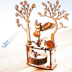 Peut inclure: Une figurine en bois représentant un hibou et un hérisson, avec des ailes déployées, perchée sur une petite table entre deux arbres stylisés. La figurine est en bois brun clair avec des détails complexes.