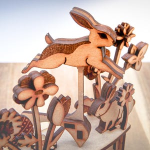 May include: Wooden Easter decoration featuring a leaping rabbit and various flowers. The laser-cut design showcases intricate details and a natural wood finish. The scene includes a bunny, tulips, and sunflowers, creating a spring theme.