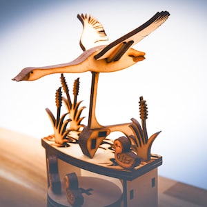 Puede incluir: Escultura de madera de un ganso volando con las alas extendidas, hecha de madera marrón claro. La escultura incluye juncos y un patito, todo sobre una base de madera. El diseño es intrincado y detallado.