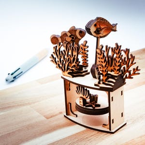 Op de afbeelding: Een lasergesneden houten aquariumscène met vissen, koraal en ander zeeleven. De miniatuurdisplay is gemaakt van lichtgekleurd hout en bevat een gedetailleerde basis met een rond platform.