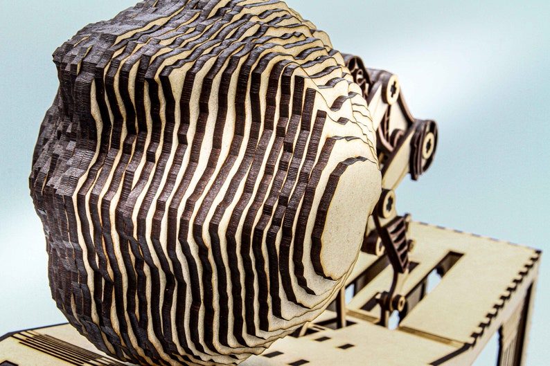 May include: A wooden 3D model of a head with a layered design. The head is made of multiple thin pieces of wood that are stacked on top of each other. The wood is a light brown color with dark brown lines.