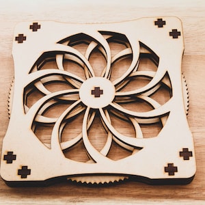 May include: A wooden, laser-cut, geometric design with a circular pattern and a gear-like edge. The design is made of light brown wood with dark brown accents.
