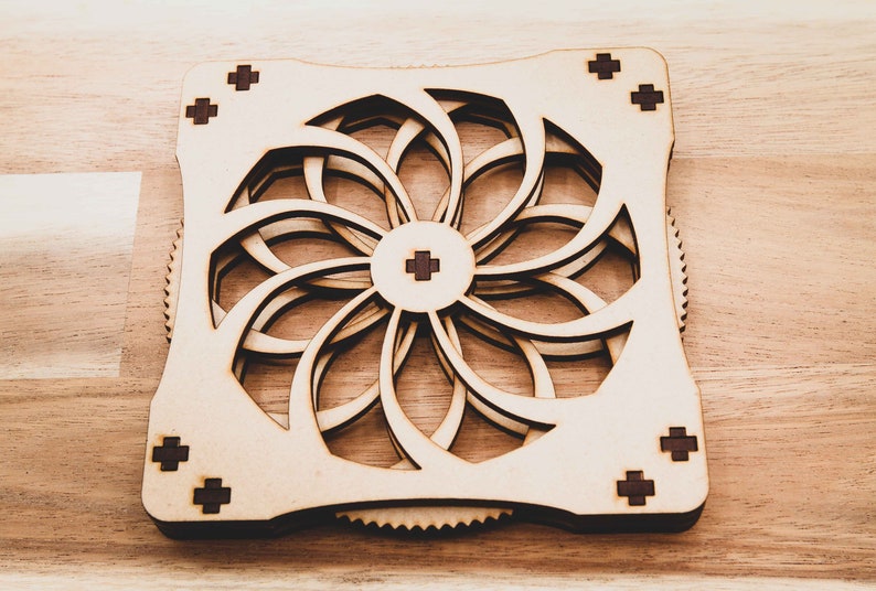 May include: A wooden, laser-cut, geometric design with a circular pattern and a gear-like edge. The design is made of light brown wood with dark brown accents.