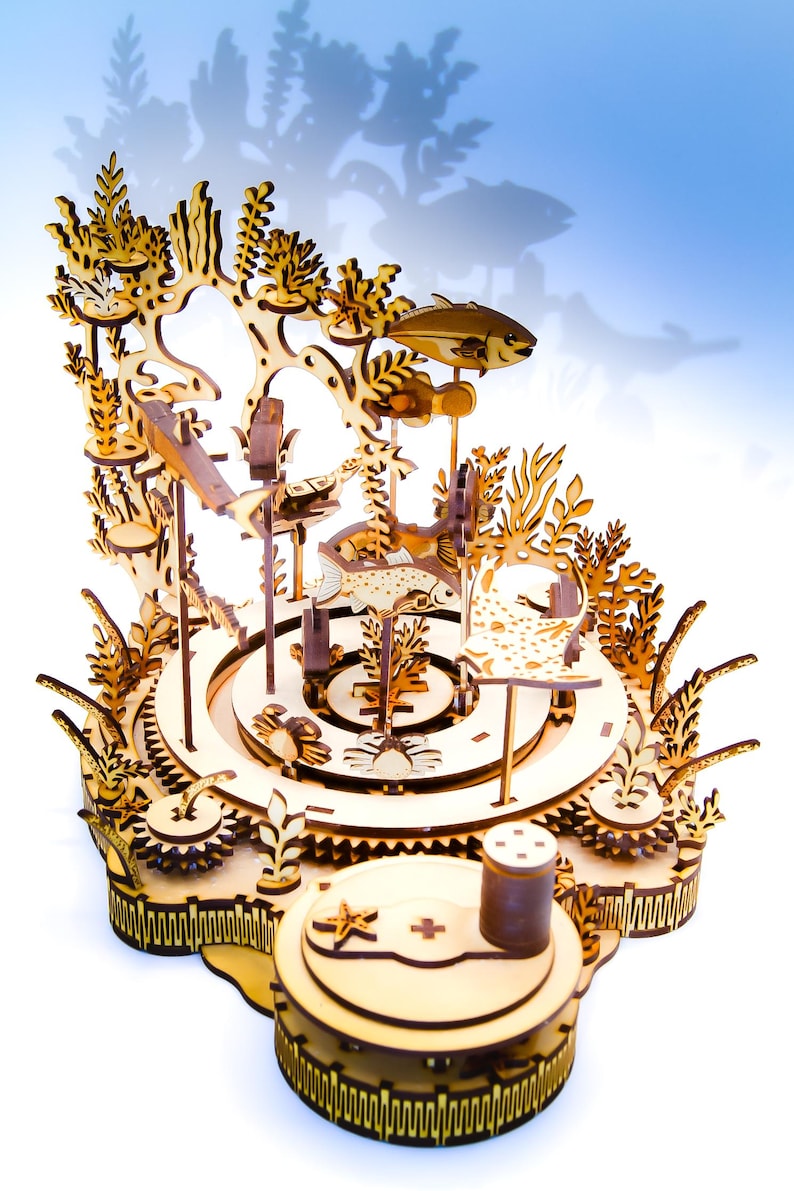 May include: A wooden, laser-cut model of an underwater scene, featuring fish, coral, and other marine life. The model has a circular base with gears and a detailed, layered design. The natural wood color is prominent.