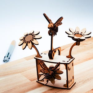 Può includere: Un elemento decorativo in legno con un design a forma di ape e fiori. Il pezzo include una piattaforma centrale con una figura di ape e due fiori. I fiori e le api sono in legno marrone chiaro, con dettagli intagliati.