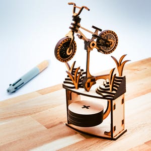 May include: A detailed wooden bicycle model, complete with spokes and handlebars, sits on a decorative stand. The stand has a layered design with cut-out grass and a circular base. The model is crafted from light brown wood.