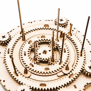 May include: A wooden gear mechanism puzzle with multiple interlocking gears, wooden dowels, and a central circular design. The puzzle is made of light-colored wood and features intricate details, including small wooden pegs and cut-out patterns.