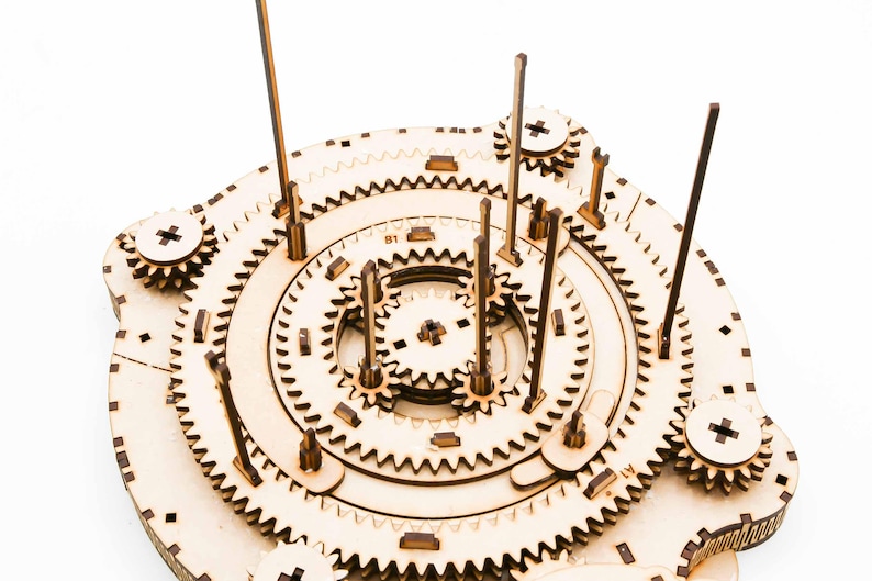May include: A wooden gear mechanism puzzle with multiple interlocking gears, wooden dowels, and a central circular design. The puzzle is made of light-colored wood and features intricate details, including small wooden pegs and cut-out patterns.