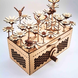 May include: A wooden music box featuring laser-cut daisies and bees. The box has a honeycomb pattern on the sides and a crank on the side. The daisies and bees are arranged on top of the box.