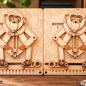 Könnte beinhalten: Zwei helle Holztafeln mit lasergeschnittenen Affenfiguren mit Fliegen. Jede Tafel zeigt Bananenillustrationen, den Text BANANA + oder BANANA X, ein nummeriertes Raster und einen Schiebemechanismus an der Basis mit den Zahlen 1 bis 12.