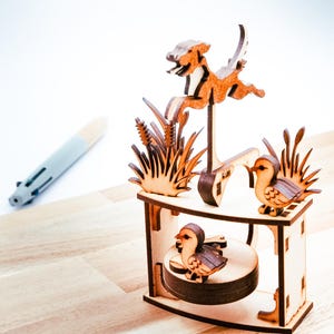 Puede incluir: Escultura cinética de madera que representa una escena de caza con un perro, patos y juncos. La escultura está hecha de madera de color marrón claro y tiene un diseño detallado y en capas. Los patos están posados en una plataforma.