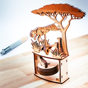 Puede incluir: Diorama de madera cortada con láser que representa una escena de sabana. Un guepardo salta sobre un árbol, con otros animales y follaje. El diorama está sobre una base rectangular. La madera es de color marrón claro natural.