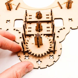May include: A partially assembled wooden gear clock kit. The unfinished wood pieces are intricately cut and designed to interlock, featuring gears, supports, and decorative elements. The clock is being assembled by hand.