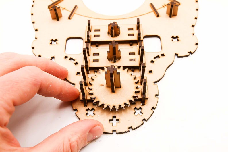 May include: A partially assembled wooden gear clock kit. The unfinished wood pieces are intricately cut and designed to interlock, featuring gears, supports, and decorative elements. The clock is being assembled by hand.