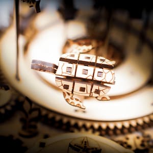 May include: A wooden turtle puzzle with a geometric design, suspended in front of a blurred background of gears and other wooden components. The turtle has a light brown color with darker brown accents.