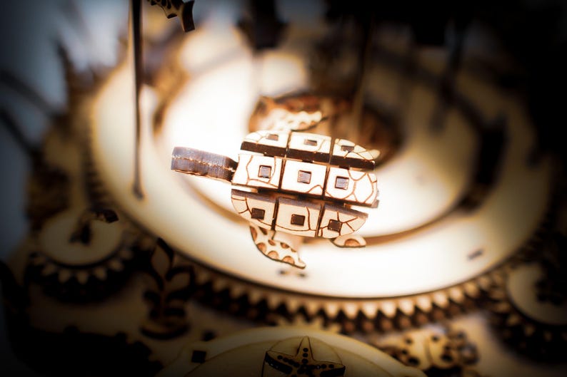 May include: A wooden turtle puzzle with a geometric design, suspended in front of a blurred background of gears and other wooden components. The turtle has a light brown color with darker brown accents.