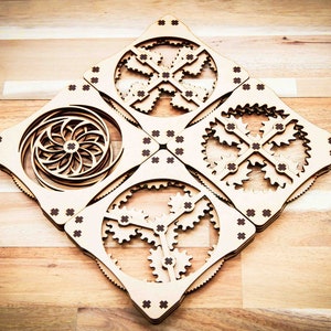 Può includere: Quattro piastrelle quadrate in legno con intricati disegni tagliati al laser. Ogni piastrella presenta un motivo diverso, tra cui ingranaggi, una spirale e un design geometrico. Le piastrelle sono collegate tra loro da piccoli perni metallici.