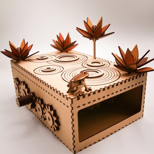 Op de afbeelding: Een houten doos met een kikker, tandwielen en lotusbloemen. De doos is gemaakt van multiplex en heeft een bruine kleur. De kikker zit op een rond platform met concentrische cirkels. De tandwielen zijn zichtbaar aan de zijkant van de doos. De lotusbloemen zijn van hout en hebben een roodbruine kleur.