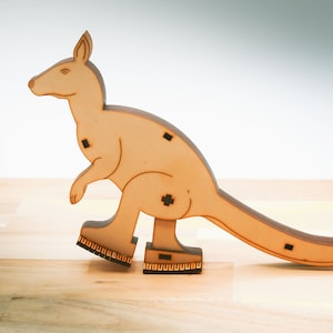 Può includere: Un giocattolo di legno a forma di canguro con una finitura marrone. Il giocattolo è in piedi su una superficie di legno e ha una lunga coda.