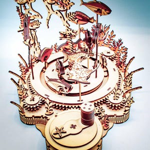 May include: A wooden, laser-cut model of an underwater scene, featuring fish, coral, and other aquatic elements. The model is constructed from light-colored wood and has intricate details, including gears and a base with a starfish design.