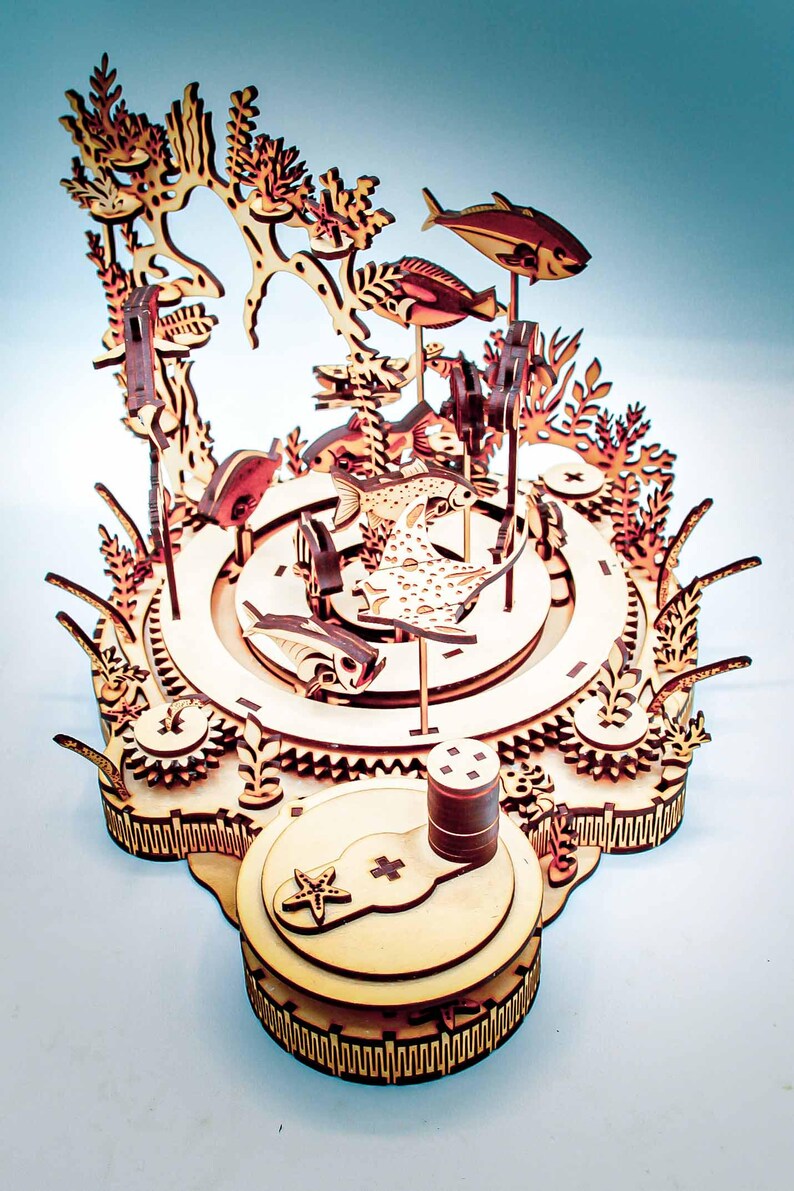 May include: A wooden, laser-cut model of an underwater scene, featuring fish, coral, and other aquatic elements. The model is constructed from light-colored wood and has intricate details, including gears and a base with a starfish design.