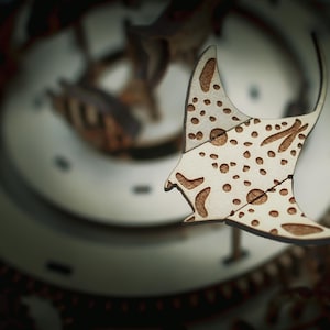 May include: A wooden manta ray cutout with a light brown finish and darker brown spots and markings. The manta ray is part of a larger wooden structure, possibly a clock or decorative piece. The background is blurred, but appears to be a complex wooden mechanism.