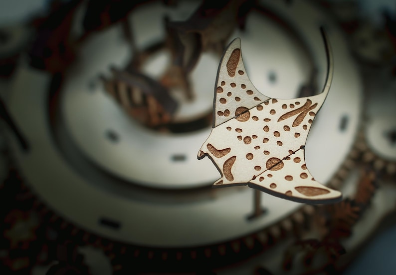 May include: A wooden manta ray cutout with a light brown finish and darker brown spots and markings. The manta ray is part of a larger wooden structure, possibly a clock or decorative piece. The background is blurred, but appears to be a complex wooden mechanism.