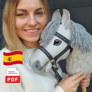 Puede incluir: Un caballo de palo con cabeza de felpa gris claro, hocico gris oscuro y una crin gruesa de lana gris. Presenta una brida gris oscuro con costuras decorativas blancas y un clip de metal, montado sobre un palo de madera con bordados.