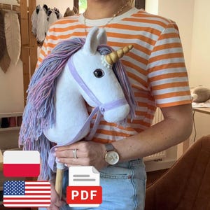 May include: A white plush unicorn hobby horse featuring a gold spiral horn, a long mane made of multi-colored pastel yarn, and a light purple fabric bridle. The unicorn head is attached to a wooden stick handle for holding and play.