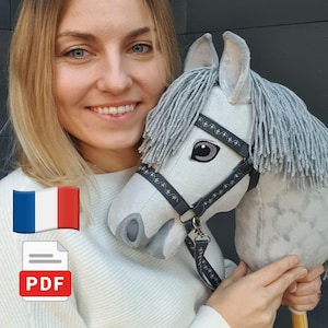 Può includere: Un cavallo giocattolo su bastone di colore grigio chiaro in tessuto morbido, dotato di una briglia grigio scuro con cuciture decorative bianche, una folta criniera in lana grigia e narici grigio scuro. La testa è montata su un manico in legno.
