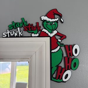 Könnte beinhalten: Ein dekoratives Tür-Eckschild mit einer grünen Figur in roter Weihnachtsmannmütze und Anzug. Der Text "stank stunk stink" steht in Grün und Rot über der Figur, während der rot-weiße Text "HO HO HO" unter der Figur angebracht ist.