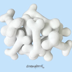 May include: A pile of white, plush bone-shaped toys against a light blue background. The toys are soft and rounded, with a three-lobed shape at each end. The logo "dreamyferret.co" is visible in the bottom right corner.