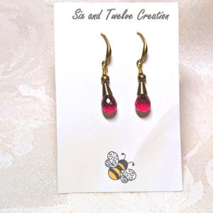 May include: A pair of dangle earrings with gold-colored hooks and accents. Each earring features a faceted, teardrop-shaped, ruby-red crystal. The earrings are displayed on a white card with the text "Six and Twelve Creation" and a bee illustration.