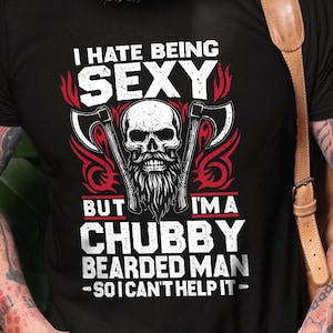 Puede incluir: Camiseta negra con el texto "I HATE BEING SEXY BUT I'MA CHUBBY BEARDED MAN - SO I CAN'T HELP IT" y un gráfico de calavera con hachas. La camiseta la lleva una persona con barba blanca y tatuajes.