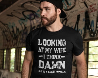 以下が含まれることがあります： 「Looking at my wife - I think - DAMN She is a lucky woman」と白文字で書かれた黒いTシャツ。