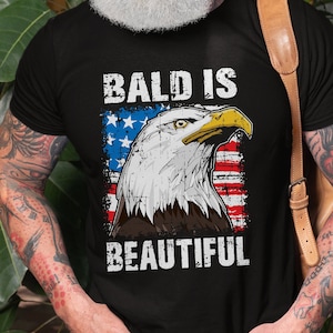 May include: A black t-shirt with a distressed American flag design and the text "Bald is Beautiful" in white lettering. An eagle with a distressed look is centered on the flag.