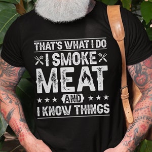 May include: A black t-shirt with a white graphic that says "That's what I do I smoke meat and I know things". The graphic includes a stylized image of a flame and two crossed grilling forks.