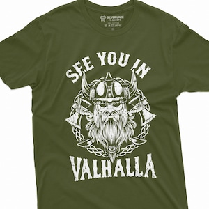 May include: Olive green t-shirt with a white graphic of a Viking warrior wearing a horned helmet, with the text "SEE YOU IN VALHALLA". The design includes axes and a braided border. The shirt is from Silver Lake T-Shirts.