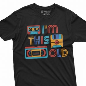 May include: Black t-shirt with a retro design featuring a cassette tape, floppy disk, and VHS tape. The text "I'm This Old" is printed on the shirt.