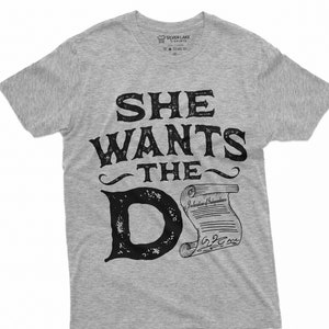 Puede incluir: Camiseta gris con un gráfico negro que dice "She wants the D" con un rollo que dice "Declaración de Independencia" debajo de la "D".