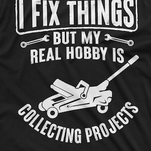 May include: A black t-shirt with white text that reads "I fix things but my real hobby is collecting projects". A white silhouette of a car jack is below the text.