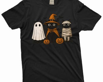 Pode incluir: T-shirt preto com um design de Halloween. O gráfico mostra três gatos fantasiados: um fantasma, uma bruxa e uma múmia. Cada gato segura uma abóbora. As cores utilizadas são preto, branco, laranja e bege.
