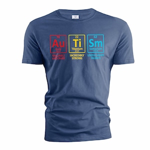 Puede incluir: Una camiseta azul marino con un gráfico de tres casillas de elementos químicos. Las casillas están etiquetadas como "Au", "Ti" y "Sm" y representan oro, titanio y samario. El texto debajo de las casillas dice "Au Ti Sm", "Oro", "Titanio", "Samario", "Variable y raro", "Increíblemente fuerte" y "Talentos útiles muchos".