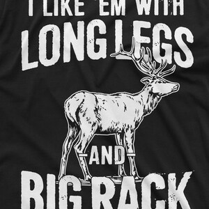 May include: A black t-shirt with white text that reads "I like 'em with long legs and big rack". A white silhouette of a deer with large antlers is in the center of the shirt.