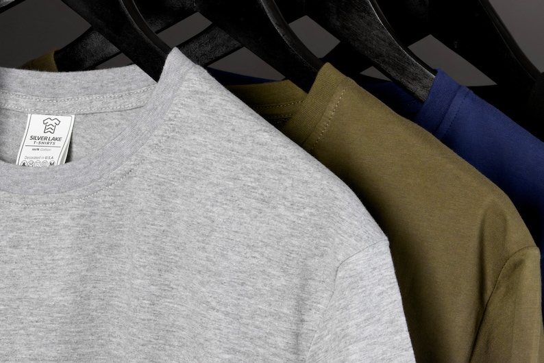 May include: Three t-shirts hanging on hangers. The t-shirts are gray, olive green, and navy blue. The gray t-shirt has a small white tag with the text "Silver Lake T-Shirts" and a graphic of a chef's hat.