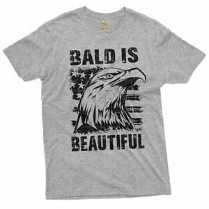 May include: Gray t-shirt with a distressed black graphic of an eagle's head with an American flag behind it. The text "Bald is Beautiful" is printed below the eagle.